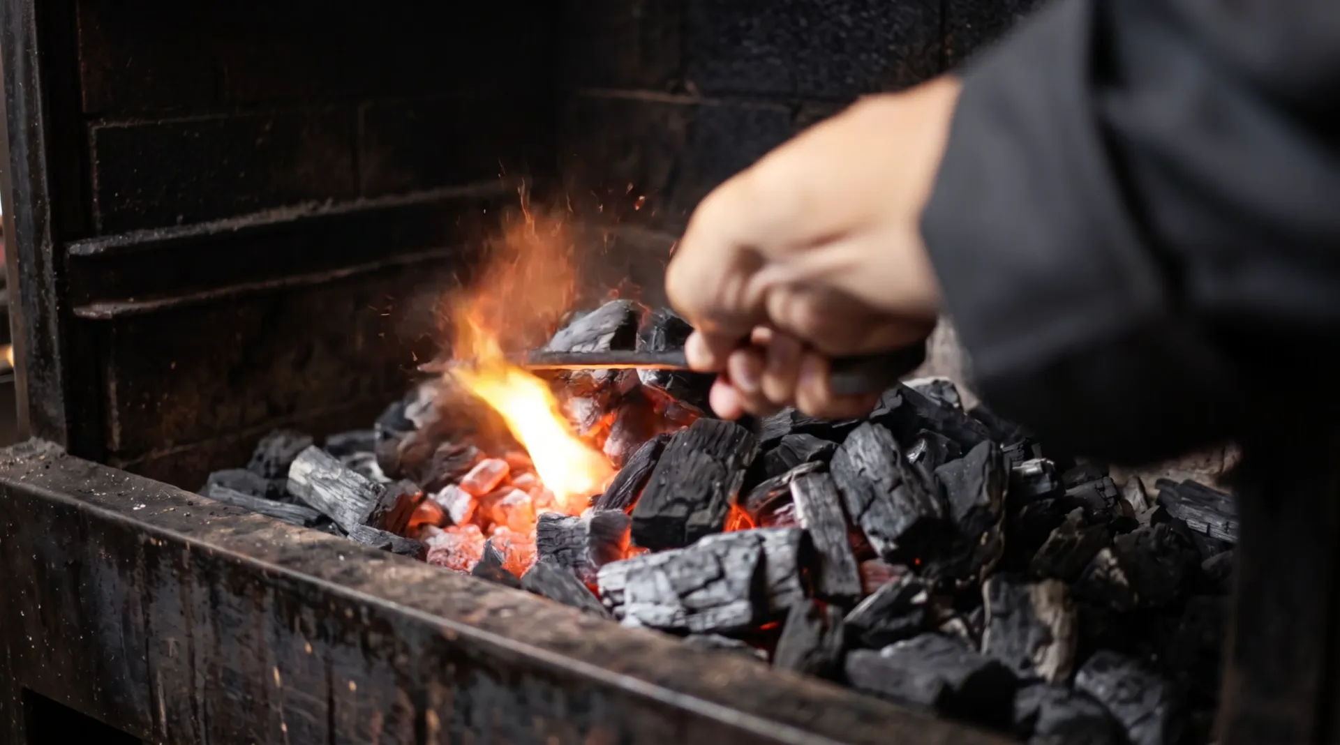 A hand rakes a bed of charcoal with an iron poker: the coals glow and a flame jumps up with sparks