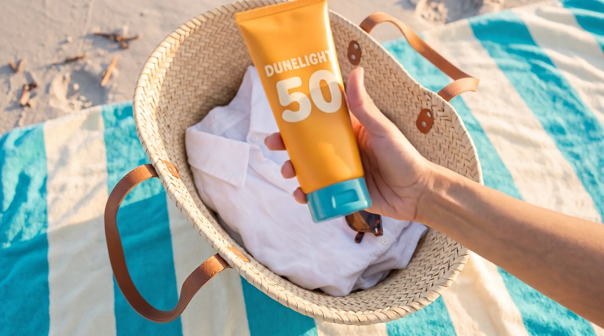 A hand lifts an orange tube with the word Dunelight and a large 50 out of an open straw bag on a striped towel