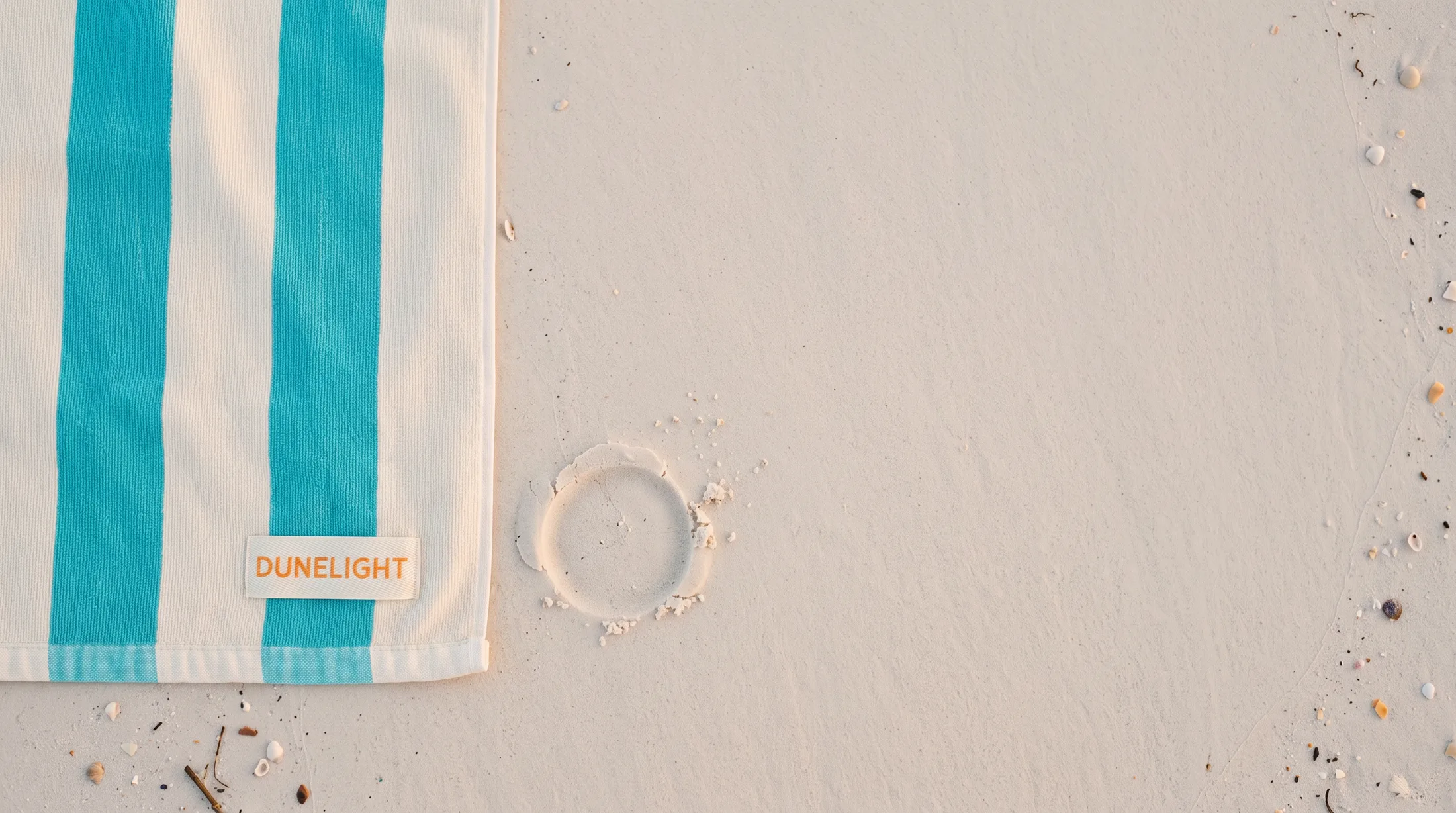 The corner of a striped beach towel on pale sand, a woven label with the word Dunelight sewn into its hem; beside it the round print of the tube's cap in the sand