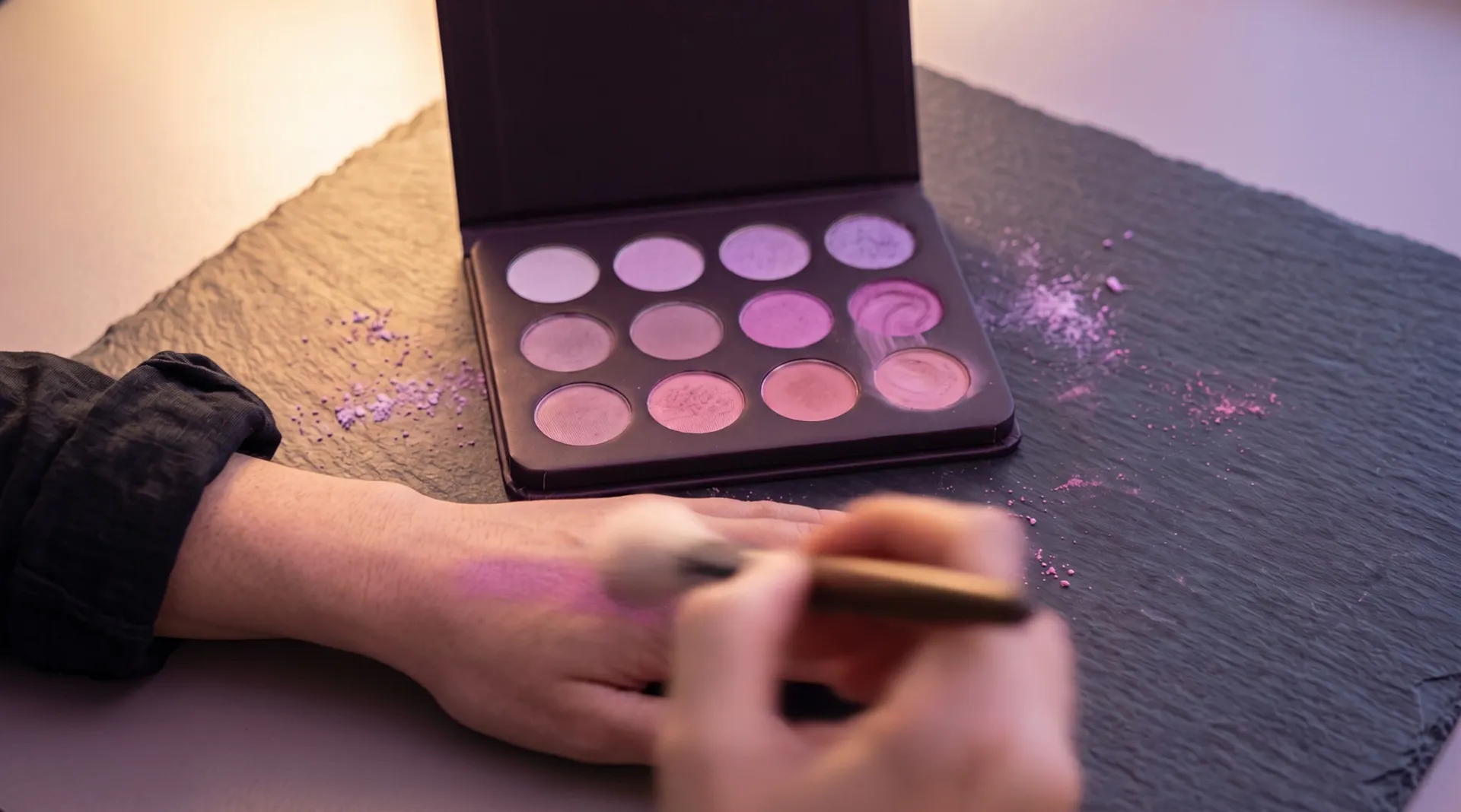 A soft brush draws a stroke of orchid pink across the back of a hand; the open palette lies beside it