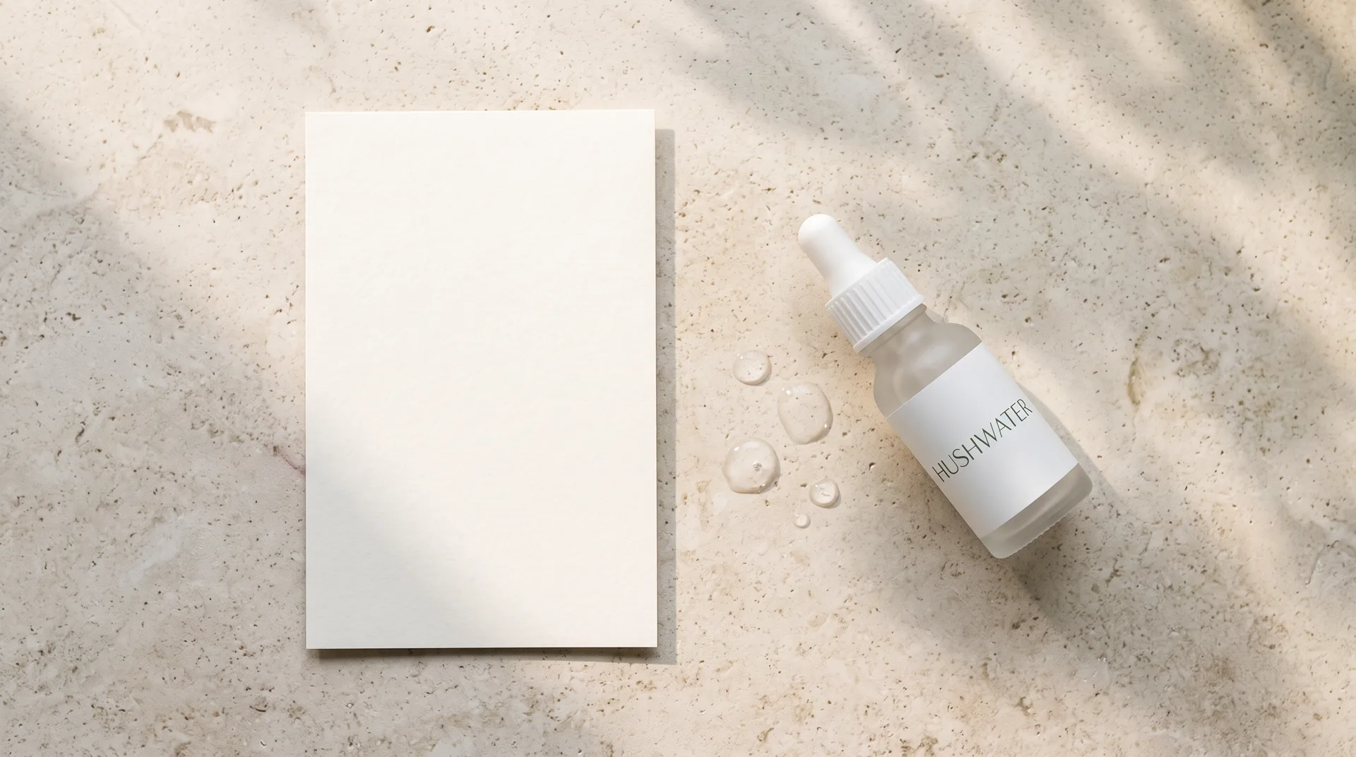 A sheet of cream label paper on pale stone in morning light; beside it the Hushwater dropper bottle lies on its side among a few drops of water