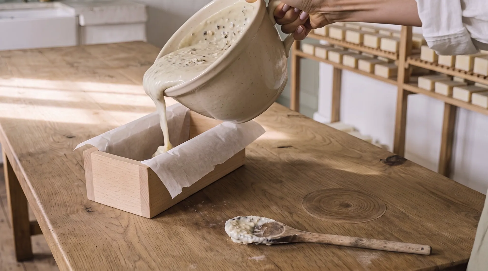 A hand tilts the stoneware bowl: thick cream soap batter runs from its spout into a wooden mould lined with parchment