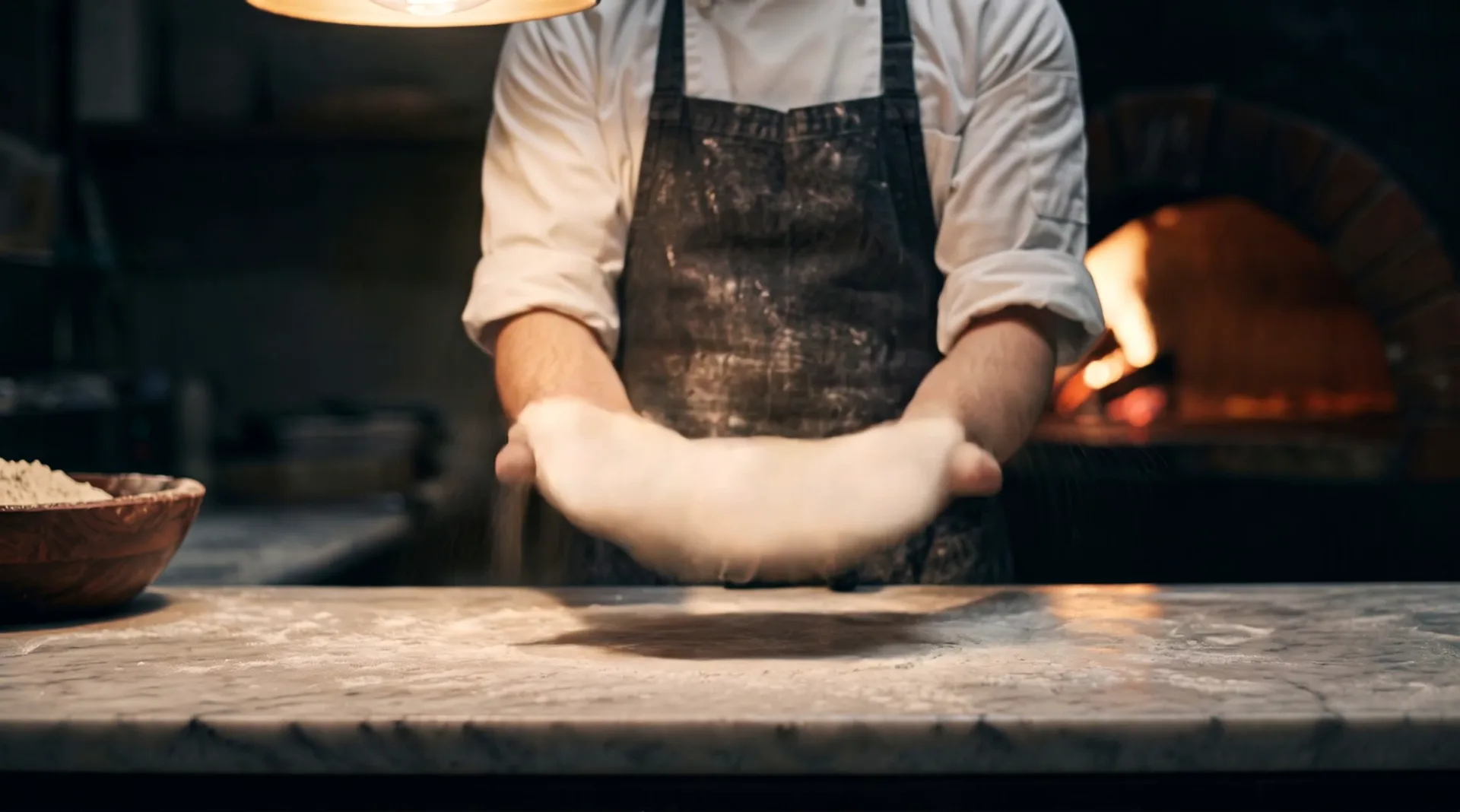 A pizza chef stretches a round of dough over his fists above a floured marble counter, in the beam of a lamp