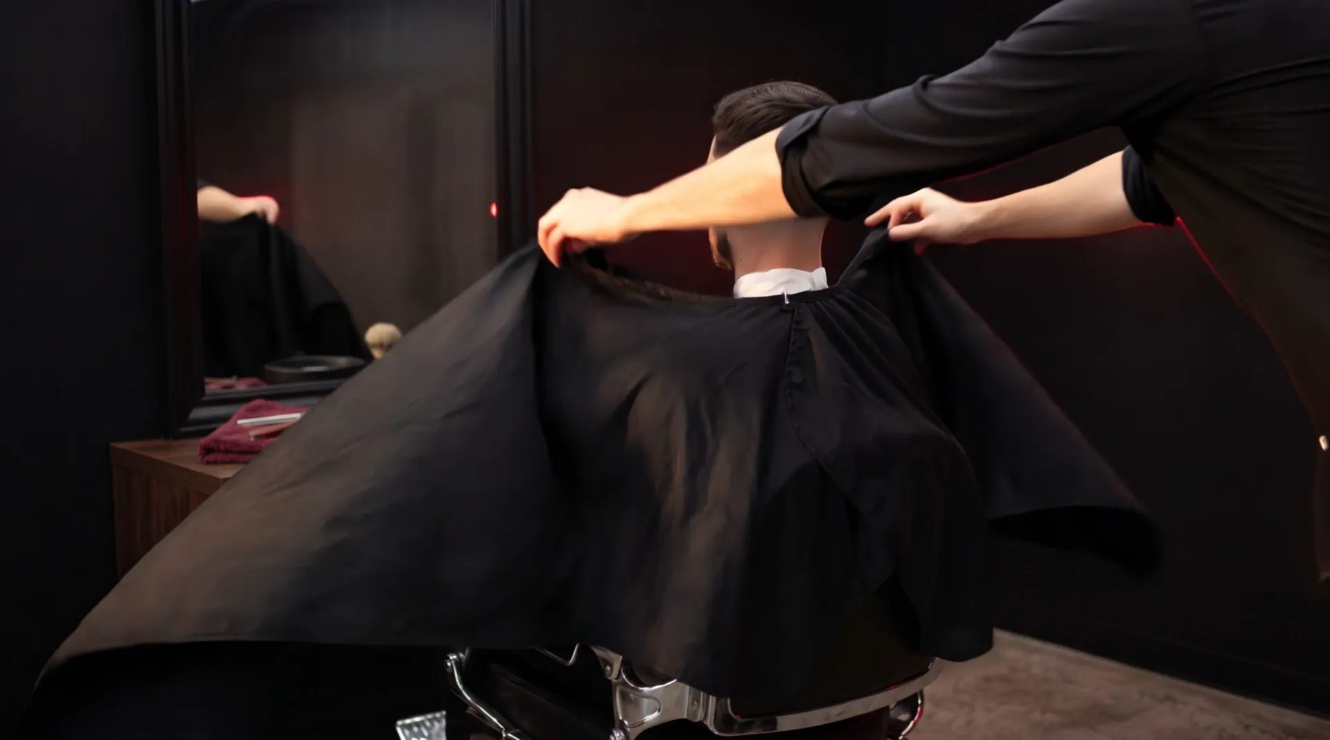 A man in a barber chair seen from behind; two hands draw a black cape round his shoulders