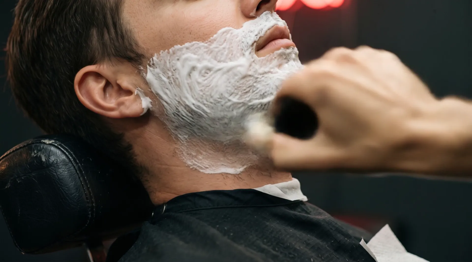 A shaving brush paints white lather onto the cheek and jaw of a man leaning back in the chair