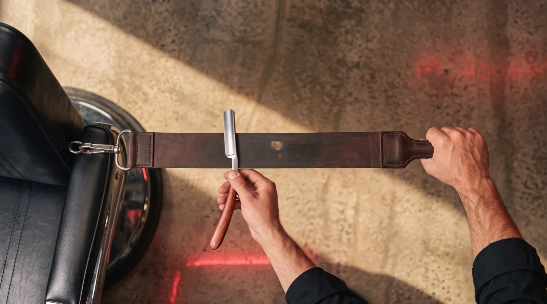 Seen from above: one hand pulls a leather strop taut from the side of the chair, the other draws an open razor along it