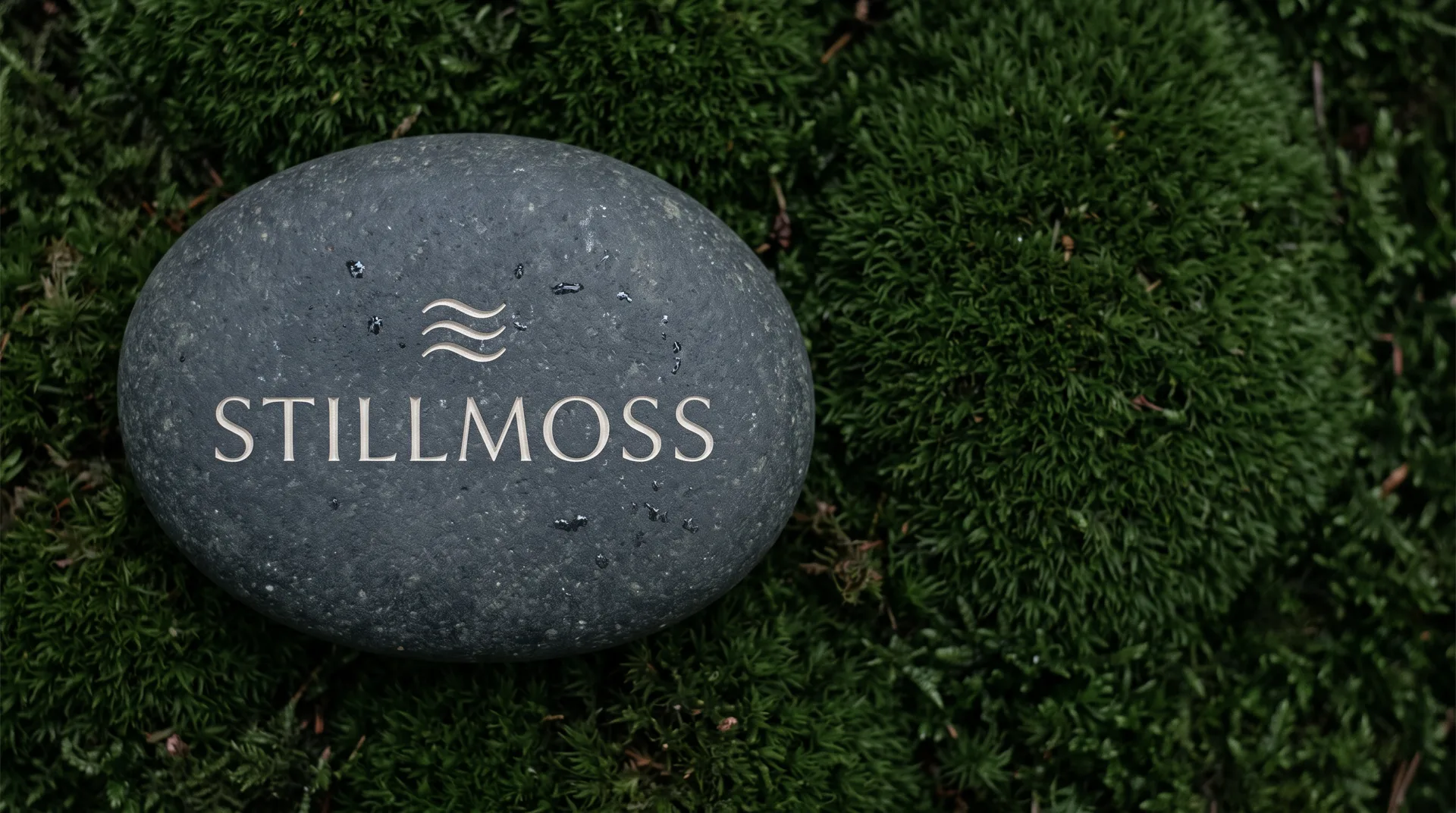 A smooth dark stone lying on green moss; three wavy lines and the name Stillmoss are carved into it