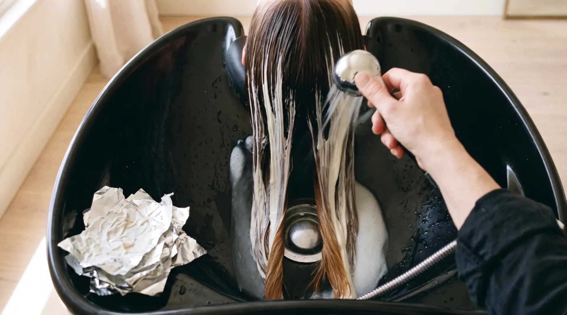 Seen from above a black basin: a hand moves a shower over long hair; the cream runs off in milky water and the ends come out the colour of caramel