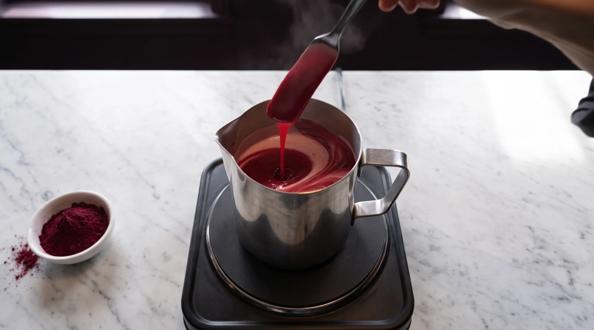 A hand lifts a spatula out of a steel pitcher on a hot plate: thick cherry-red lipstick mass runs off the blade in a ribbon